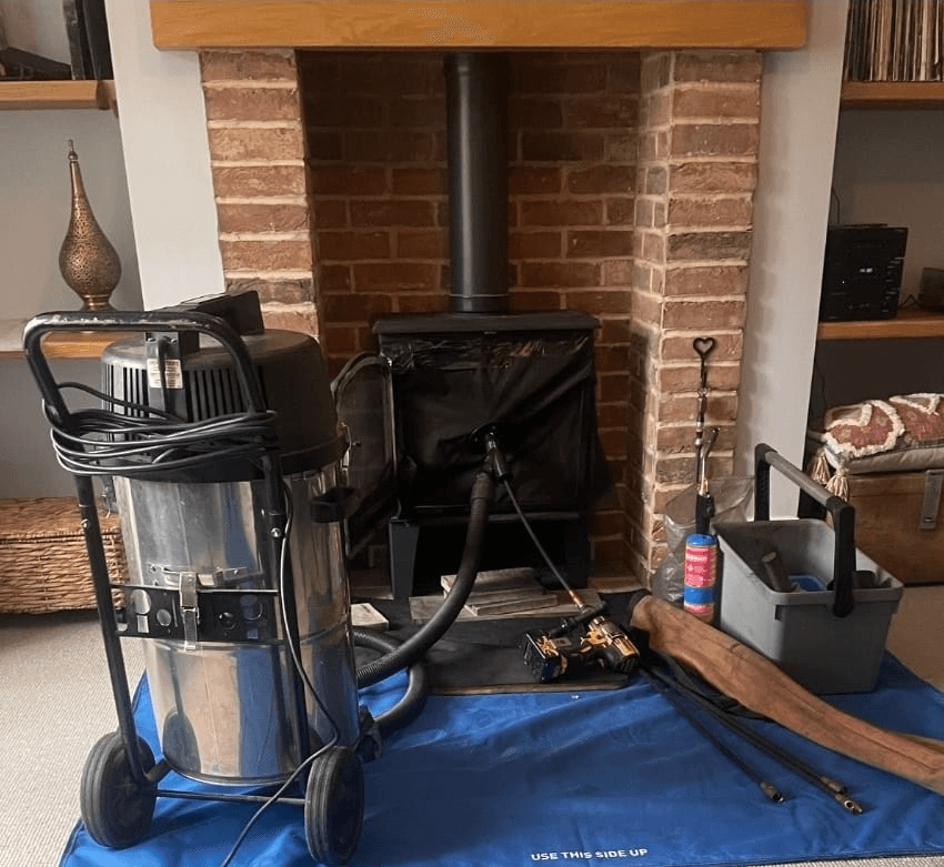 A chimney sweep's vacuum, rods and tool caddy set up on dust sheets beside a brick inglenook fireplace.