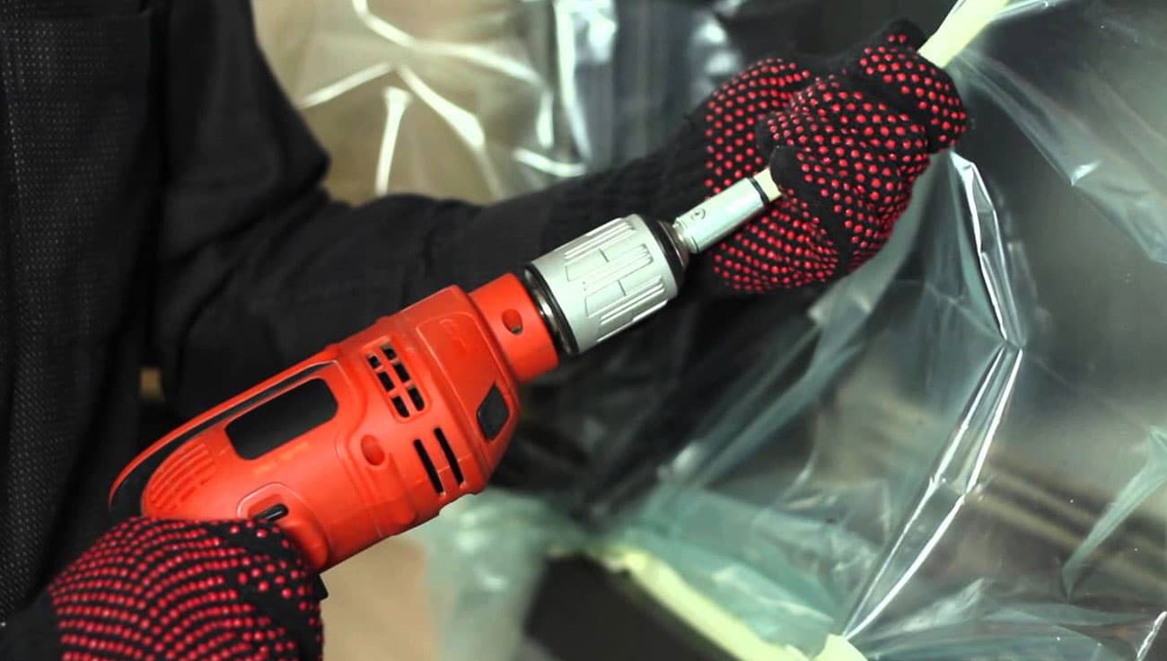Gloved hands using a rotary power-sweeping adaptor on a drill, with protective sheeting in place.